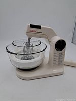 Off-white vintage Sunbeam Mixmaster stand mixer with a glass mixing bowl and two beaters attached. The mixer has a large dial with speed numbers and setting labels on the side.