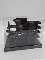 Front and diagonal view of black plastic mechanical novelty clock in clear protective case showing horizontal numbered rails and metal balls.