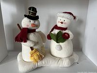 Two animated musical snowmen figures on a white snow base, male with black plaid top hat and broom, female with red Santa hat and holding green book.