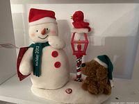 Front view of the 2013 Hallmark Jingle Pals Merry Carolers Trio showing the snowman with red buttons and green scarf, a red bird atop the candy cane-striped lighted lamp, and the brown dog with a Santa hat on a white snowy base.