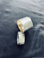 Photo displaying two antique silver napkin rings on a dark background: one round with floral engravings and one plain oval ring.