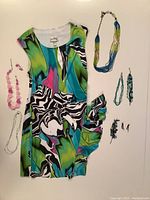 Full view of the Joseph Ribkoff multicolor slimming jersey dress with side ruffle, laid flat with jewelry arranged beside it showing multiple necklaces, bracelets, and earrings in complementary colors.