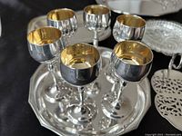 Close-up of six silver-plated liquor glasses with gold wash interior on a small silver tray, showing reflective surfaces and gold lining.