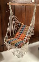 Front view of the hanging chair suspended from a wooden spreader bar showing foam cushions in striped multicolor fabric and macrame trim.