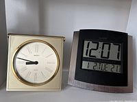 Front view of Seiko gold tone analog clock with Roman numeral dial next to La Crosse digital clock showing LCD time and date