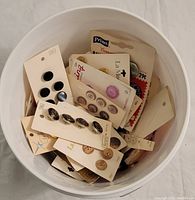 White bucket filled with mostly packaged vintage buttons on cards, black, silver, purple, and tan colors visible.