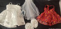 Three Build-a-Bear outfits laid out including a white wedding dress, veil, shoes, and a floral red sun dress.