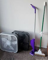View showing two box fans, white and black, alongside a mop and a Swiffer-style cleaner with purple handle.