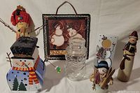 Full view of all seven snowman-themed holiday decorations against a plain background showing the slate plaque, tea light holder, glass jar, picture frame, hanging snowman figure, and two figurines.