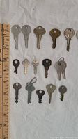 17 keys laid out on white cloth beside a 12 inch ruler for scale, showing variety of metal keys of different sizes and shapes including silver, brass and dark toned keys.