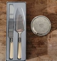 Cake cutting set including knife and pie server with ivory handles in tray, alongside a round silver coaster.