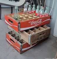 Three stacked wooden bottle crates with several vintage glass bottles inside the top crate. The top and bottom crates are Coca-Cola branded with red paint and white logos. The middle crate is a natural wood color with green "G.C.C. Beverages" lettering. Metal corner brackets are visible on the crates.