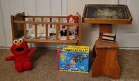 Photo showing the vintage wooden doll bed with two dolls, baby blankets, a rustic wooden school desk and bench, 1996 Tickle Me Elmo plush, and world map puzzle box on carpeted floor.