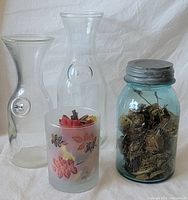 Two clear glass bottles, a blue ball jar with dried flowers inside and metal lid, and a frosted glass with faux fall leaves inside.