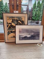Two artworks side by side on floor against window: large mountain print in multi-panel frame and smaller misty mountain landscape framed print.