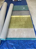 Photo showing the full length of the rug partially rolled with visible stripes in green, yellow, gray, beige, and teal on a blue tarp.