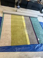 Full view of the large area rug partially rolled open, showing vertical stripes of cream, green, yellow, taupe, and blue colors. The rug is on a blue tarp in a garage setting.