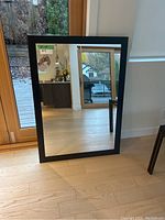 Black framed rectangular mirror leaning against a wall next to a large glass door, showing clear reflections of the interior and exterior environment.