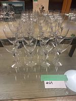 Front angle view of 22 stemmed large wine glasses arranged on a glass table, showing their tall stems and wide bowls.