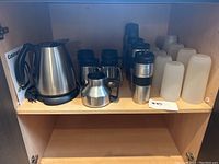 Photo showing Cuisinart kettle, travel mugs including stainless steel Starbucks cups, and white plastic cups on a cabinet shelf