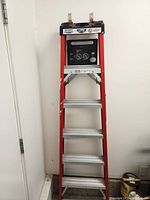 Full front view of the red 6-step ladder with aluminum steps against a white wall.