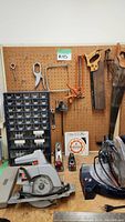 View of pegboard with hand tools, hand saws, hacksaw, hand planes, and small bins with screws and hardware. Also includes two red and silver hand planes and pliers.