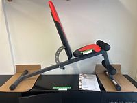 Side view of the workout bench showing the angled padded backrest, seat, and foam roller leg holders with the metal frame; manual and packaging visible beneath bench