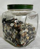 Front and side view of vintage glass jar filled with multiple buttons in neutral tones with embossed decorative pattern on jar and green screw lid.