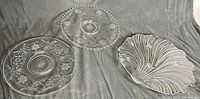 Three dessert serving plates shown together on gray fabric background. Two round plates with etched floral decorations; one with beaded rim (Cornflower), the other with starburst center. Third plate is leaf-shaped with ridged texture.