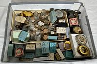 Box containing multiple small glass vials with cork stoppers and labeled boxes, displaying an assortment of watch parts and tools.