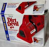 View of the Dirt Devil Plus red handheld vacuum cleaner in original box showing logos, color, and packaging details including paper bag adapter included.
