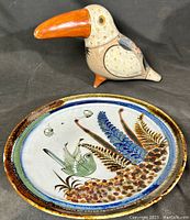 Toucan ceramic figurine with orange beak and speckled cream body next to decorative plate with painted bird and foliage design