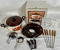 Full set including pot with lid, burner, metal stand, six fondue forks with wooden handles, three pot stands/clips, instruction sheet, and original box with pot floral image.