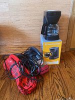 Toastmaster drip coffee maker atop original box alongside red Dirt Devil handheld vacuum and red pet hair eraser vacuum with cords and red storage bag on wooden floor.