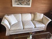 Front view of off-white 3 seater couch with two matching cushions, placed in room with hardwood floors and artwork on wall.