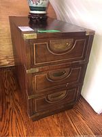 Front view showing three drawers with brass pull handles and brass corner reinforcements on top, against wooden flooring.