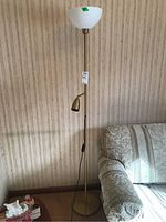 Metal floor lamp with white plastic shade and brass reading light, standing next to a couch and corner table.