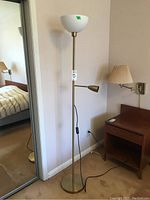 Full view of metal floor lamp with white plastic shade at top and adjustable reading light attached mid-way on pole standing near wall and next to nightstand
