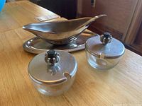 Photo showing stainless steel gravy boat with ladle on an oval serving tray and two small round glass jars with stainless steel lids and spoon slots.