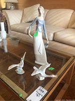 Wide shot of three Lladro porcelain figurines on glass table with beige sofa in background. Includes tall woman figure with basket and two bird figurines, one bird has glued repair.