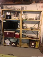 Two beige adjustable metal shelving units with four shelves each, holding kitchen appliances, coolers, glassware, and storage boxes, located in basement with visible concrete wall and pipes.