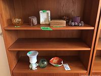 Two wooden shelves displaying various vintage glass collectibles including a milk glass vase, glass paperweight, decorative dish, trinket box, and votive holders.