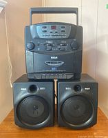 Front view of RCA BassReflex stereo center console placed on top of the two RCA BassReflex bookshelf speakers. Center console black with integrated CD player, radio tuner, cassette, and control buttons and knobs. Speakers black with visible speaker cones and bass ports.