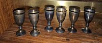 Frontal view of six tarnished stemmed liqueur goblets on wooden shelf