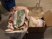 Photo showing partial box with assorted table linens including fabric placemats, napkins, and white/beige tablecloths alongside a brown stained vinyl piece.