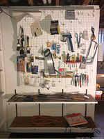Wide view of pegboard and shelving with assorted tools including handsaws, paintbrushes, and a staple gun over white pegboard with various hooks and small packages.