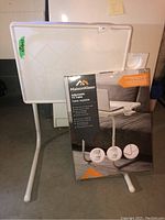 Photo showing the assembled MaisonKleen adjustable TV table next to its original packaging box, highlighting the white flat table surface and metal legs.