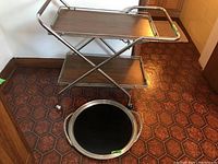 Full view of the aluminum framed folding bar cart with two wood-look shelves and a removable black surface tray with aluminum frame placed in front.