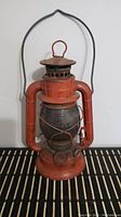 Front view of red metal Dietz lantern with wire handle and glass chimney on black and beige striped surface.