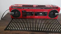 Full front view of the red Sanyo boombox on a black and beige mat, showing speakers, cassette decks, graphic equalizer, and top controls.